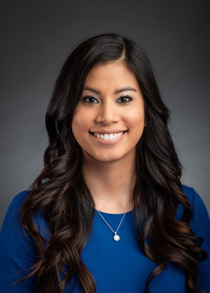 Corporate headshot of a woman with long dark hair, wearing a blue shirt, exuding professionalism and confidence.