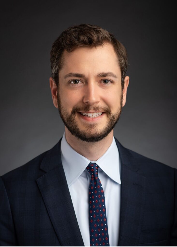 A smiling man dressed in a suit and tie, captured in a professional headshot, radiating approachability and professionalism.